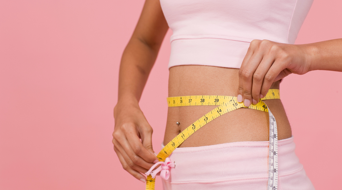 Woman in a pink crop top measuring her waist with a yellow tape measure against a pink background