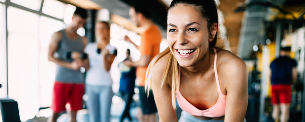 Smiling woman in a pink sports top working out in a busy gym with other people exercising behind her