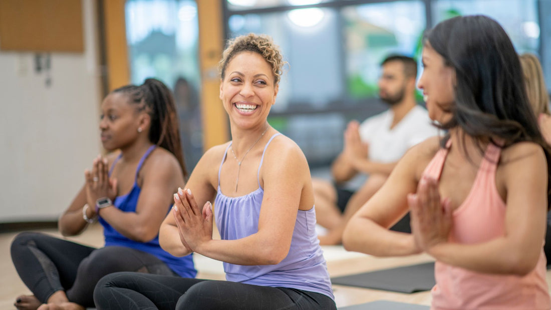Smiling adults sitting cross-legged in a group yoga class with their palms pressed together