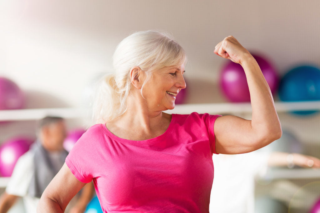 Smiling older woman in a pink T-shirt flexing her bicep in a gym
