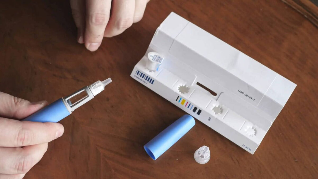 Hands assembling a blue injection pen next to a box of pen needles on a wooden table
