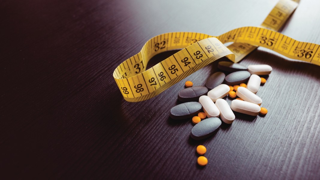 Assorted white, gray and orange pills next to a yellow measuring tape on a dark table