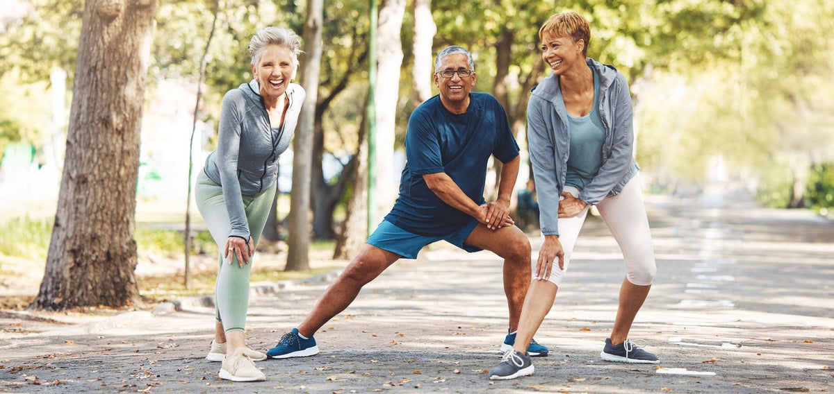 Three smiling older adults doing lunge stretches together on a tree-lined park path