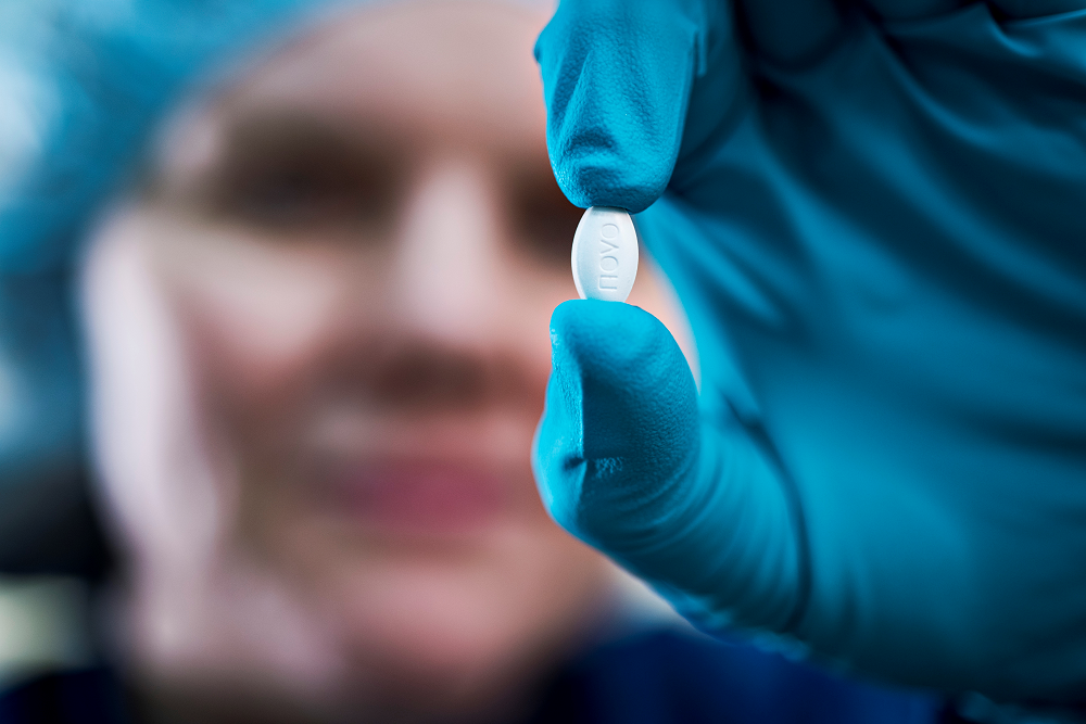 Person in blue medical gloves holding up a small white tablet, face blurred in the background