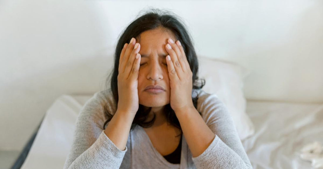 Woman sitting on a bed with her hands pressed to her face, looking tired and unwell