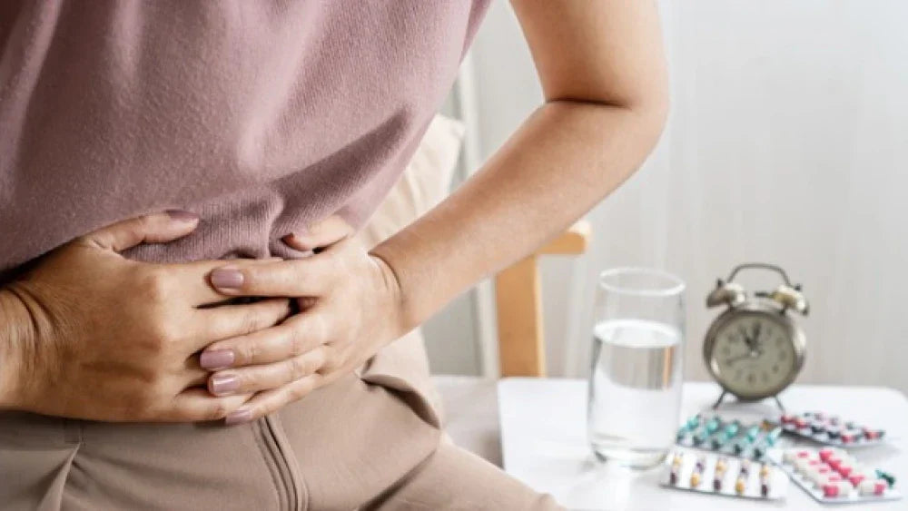 Woman holding her stomach in discomfort beside a glass of water, an alarm clock and blister packs of pills